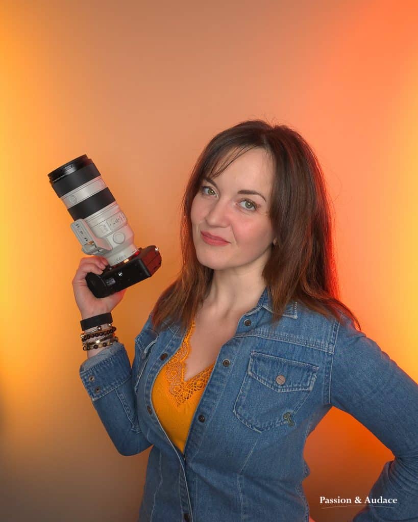 Portrait de 3/4 d'une femme, Sophie Fayet, fondatrice de Passion & Audace, tenant un appareil photo avec téléobjectif dans sa main droite sur un fond dégradé jaune orange