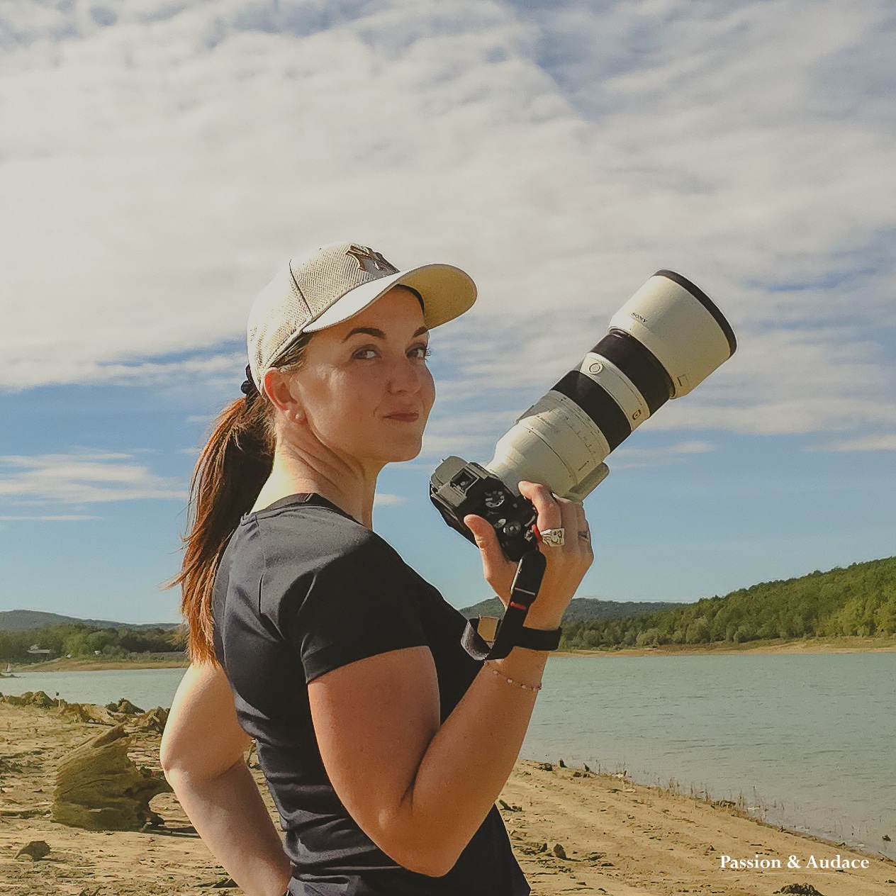 Portrait d'une femme photographe avec une casquette et tenant son téléobjectif dans la main et regardant vers le photographe sur un fond de nature et lac.