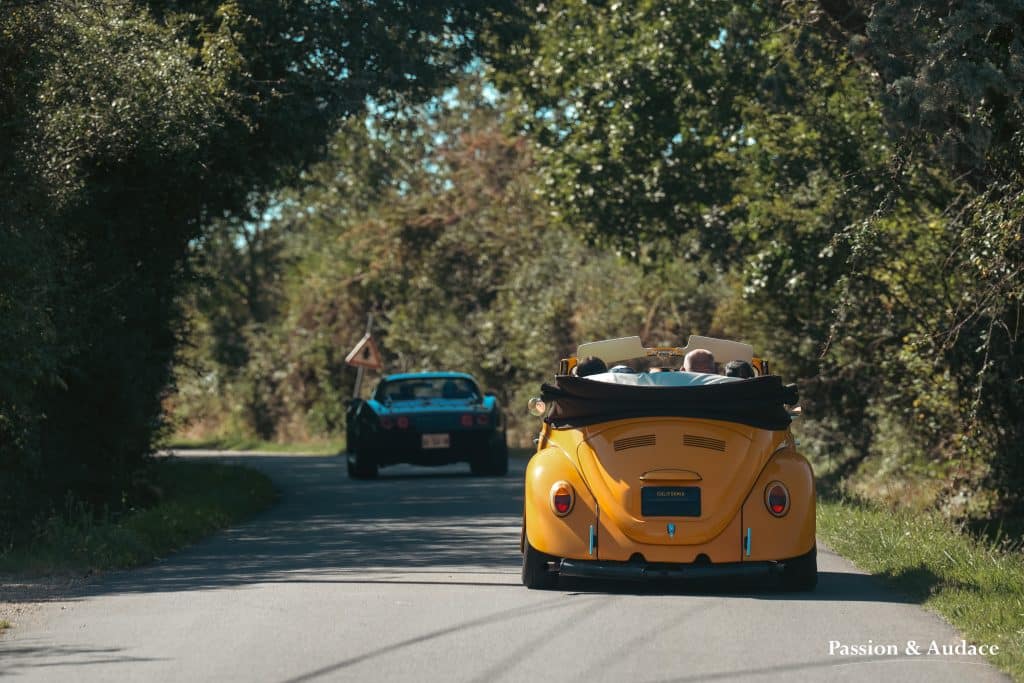 Photographie type cinématographique d'une Volkswagen Coccinelle rétro de couleur jaune orange suivant un véhicule sur la route. Image de verdure de part et d'autre se joignant au dessus des véhicules tel un pont. Photo par Passion & Audace