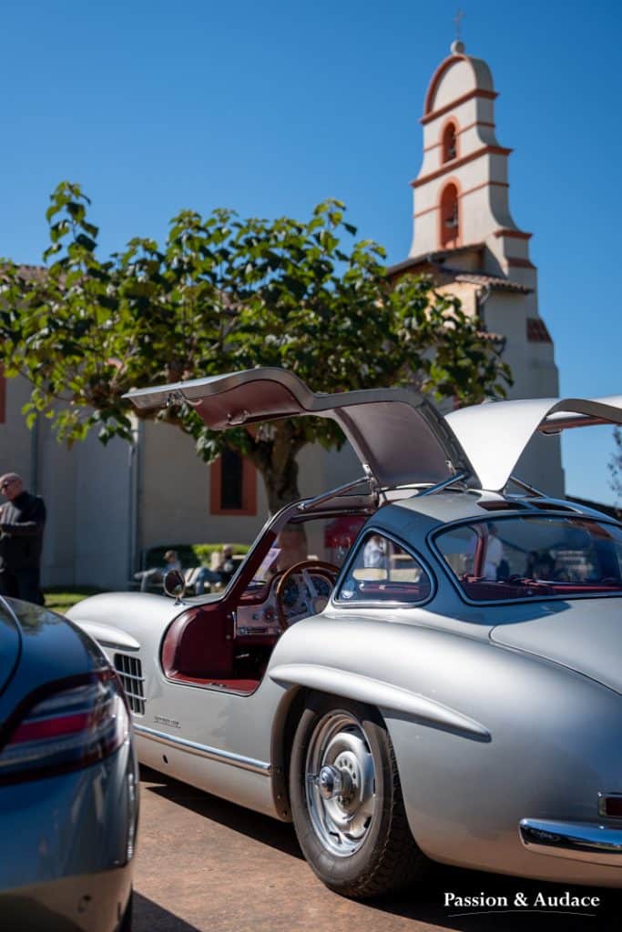 Photographie d'une Mercedes 300SL grise les portes papillon en l'air. Photo prise de 3/4 arrière avec une église à l'arrière-plan.