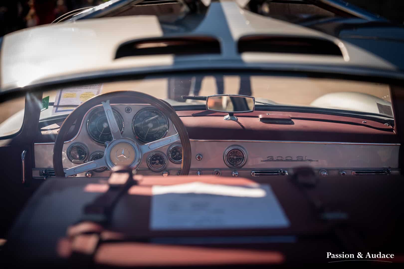Photographie du tableau de bord d'une Mercedes 300SL. Photo prise depuis la lunette arrière. Volant bois et cuir rouge.