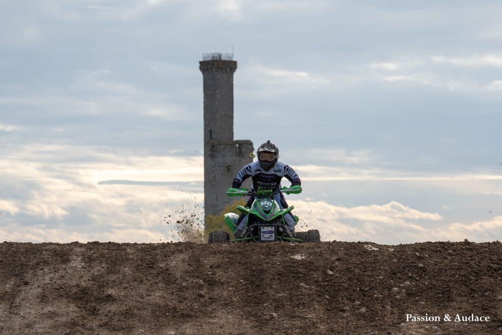 Photographie d'un pilote de quadcross venant d'atterir sur une bute de terre avec un chateau d'eau à l'arrière-plan
